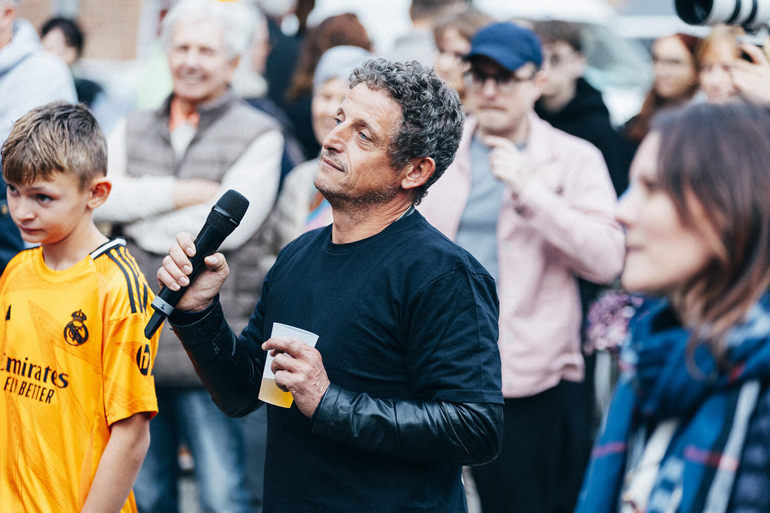 Un homme, micro à la main, s'exprime devant un public lors d'un événement. Il tient un gobelet et porte une veste noire. À gauche, un enfant en maillot de football jaune l'écoute attentivement.