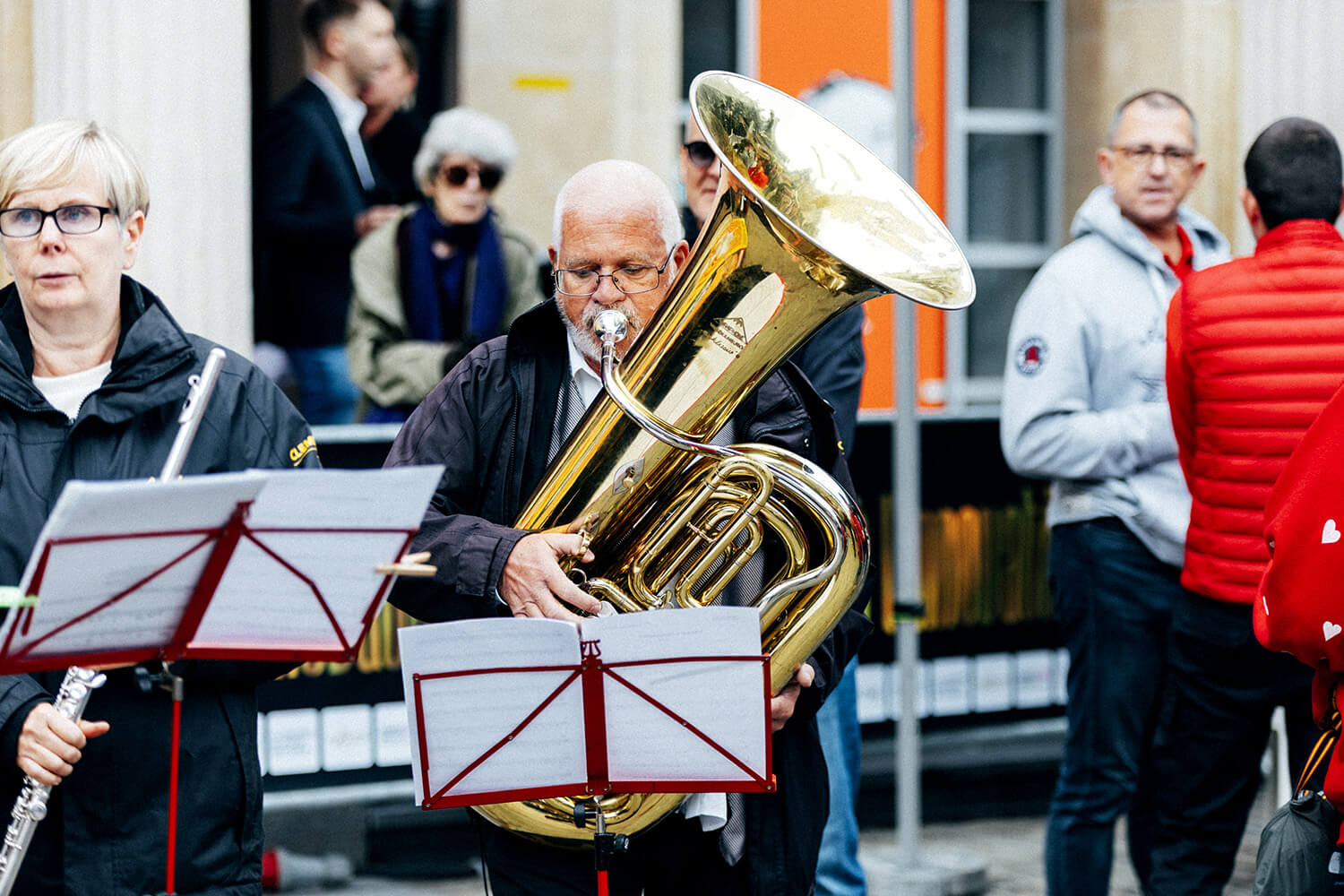 Un tubiste et une flûtiste de la fanfare d’Assesse jouent en harmonie lors d’une performance en extérieur. Les spectateurs, en arrière-plan, écoutent attentivement ce moment musical improvisé.